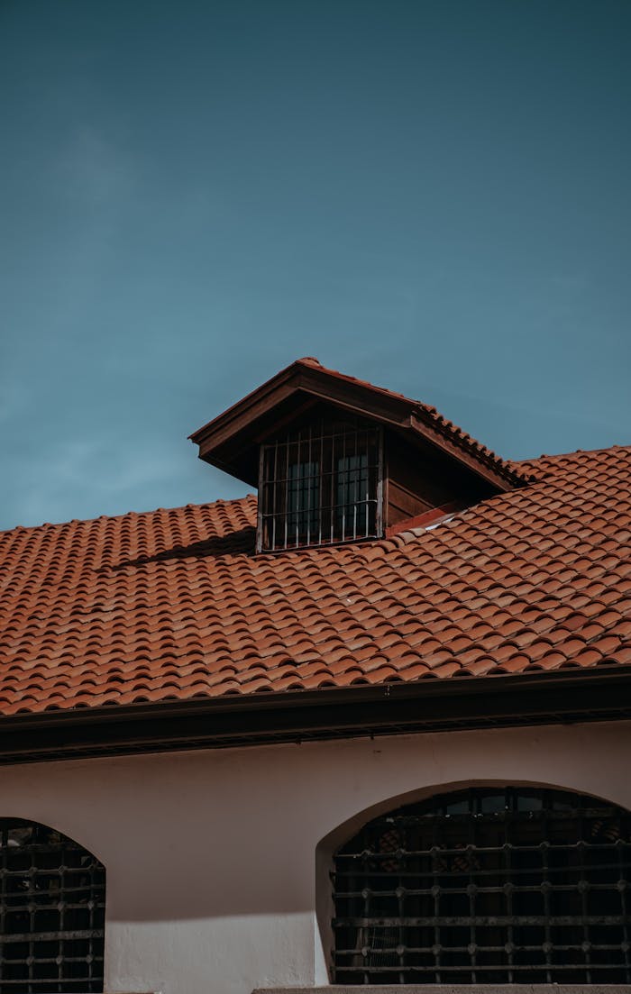 A traditional red tiled roof with an attic window under a clear blue sky, showcasing classic architecture.